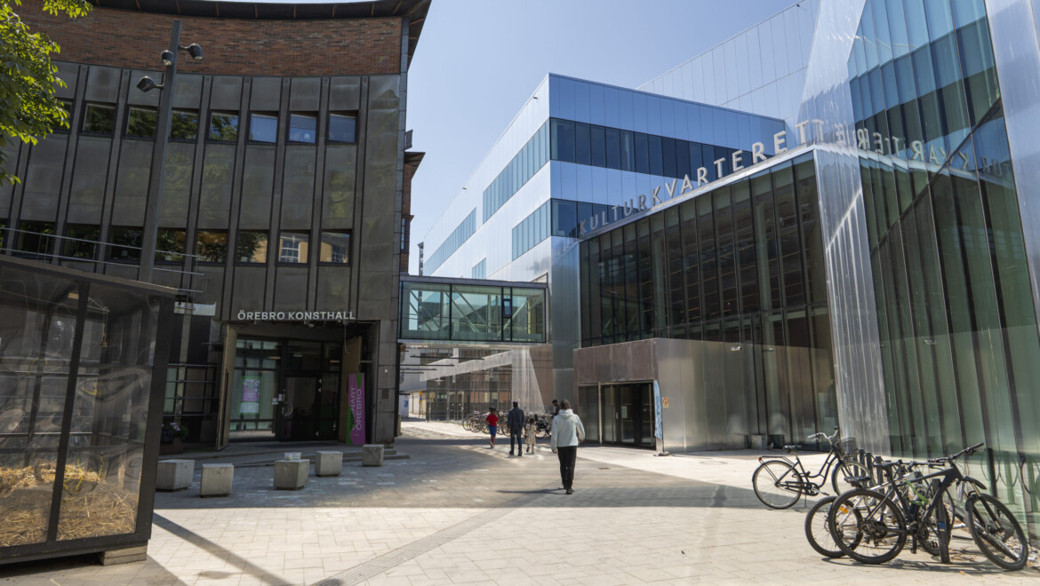 Några personer går på ett litet torg. Torget omringas av två stora byggnader, en med glasfasad och en i tegel.