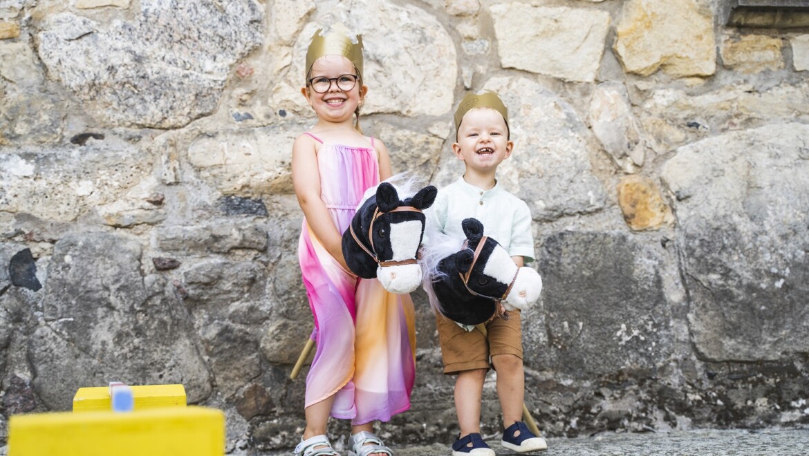 Två barn, en flicka och en pojke, står med käpphästar mot en stenvägg.