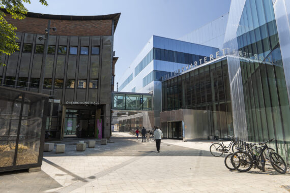 Människor går på ett torg som omringas av två stora byggnader. Den ena har en spegelfasad och texten 