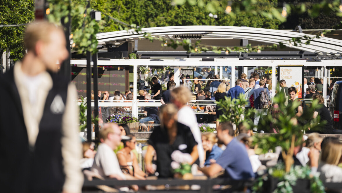 En grupp människor är påväg in på en stor uteservering. I förgrunden syns ytterligare en uteservering där många människor sitter. Det är soligt väder.