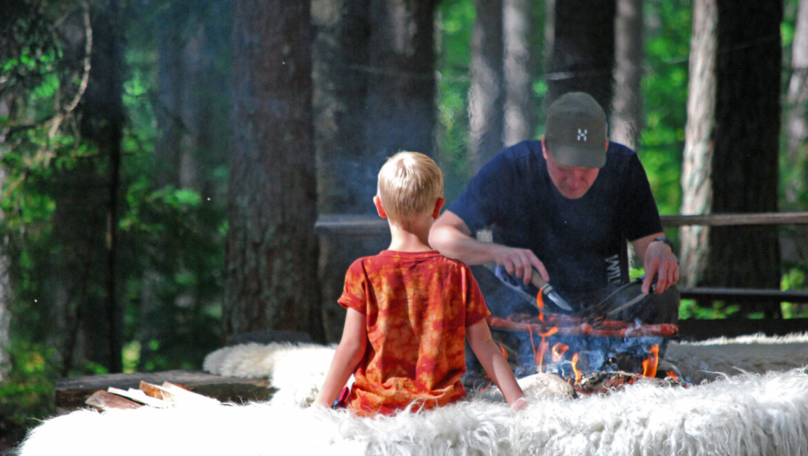 Liten pojke sitter på stor fårfäll i skogen med ryggen mot kameran mittemot en vuxen man som grillar.