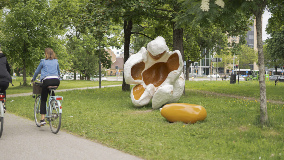 Two people are riding bicycles in a lush park. Next to the cyclists are an oversized piece of popcorn and a popcorn kernel.