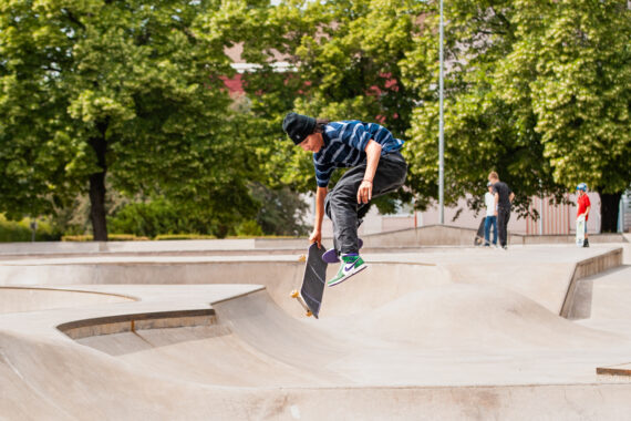 Ung man i randig tröja och mössa gör ett skateboardtrick i en betongramp på en skatepark i Örebro, med träd och andra skateboardåkare i bakgrunden.