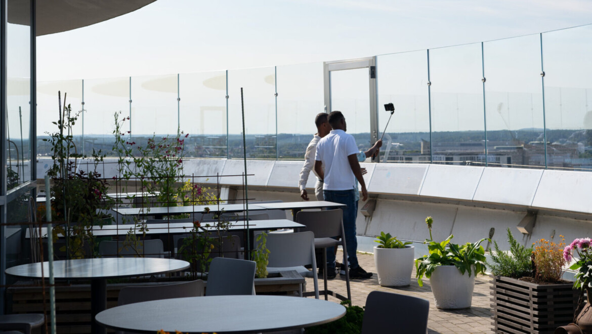 Två personer syns snett bakifrån med selfiestick på en plats med utsikt.