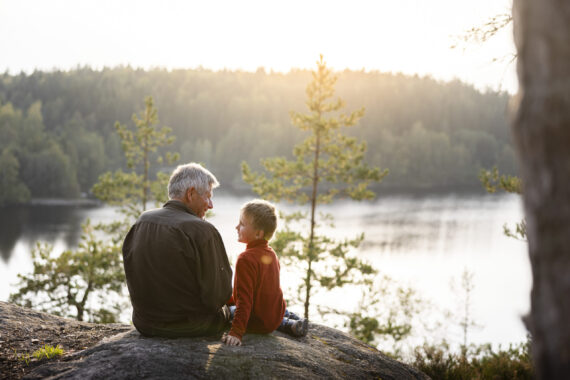 En äldre man och ett barn sitter tillsammans på en klippa med utsikt över en sjö omgiven av skog. De ser på varandra och ler i kvällsljuset.