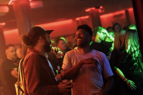 Två personer i rödbelyst nattklubbsmiljö skrattar med varandra,