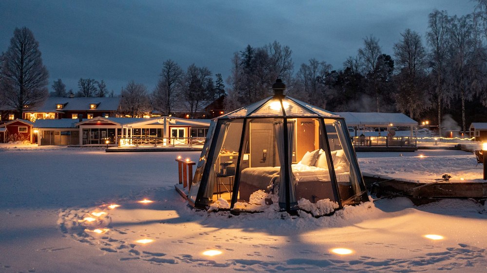 En glasigloo förtöjd vid en brygga. I igloon syns en säng. Runt omkring ligger det snö och i bakgrunden syns upplysta hus.
