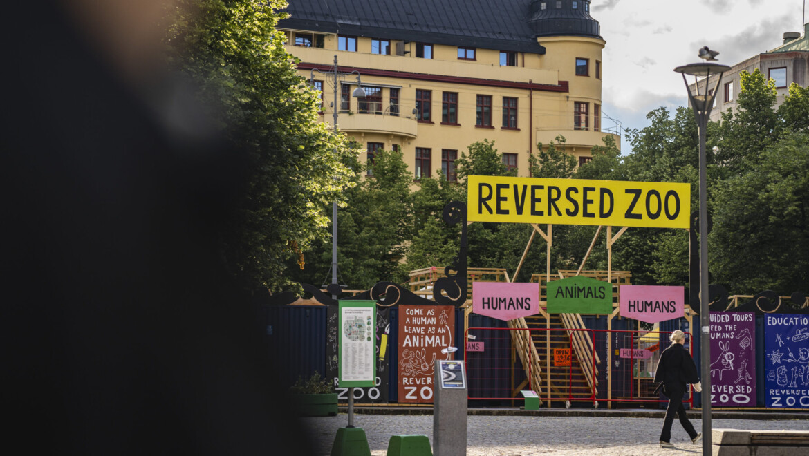 Ett stort färgglatt konstverk står placerat på ett torg. Konstverket omringas av växtlighet och höga hus.