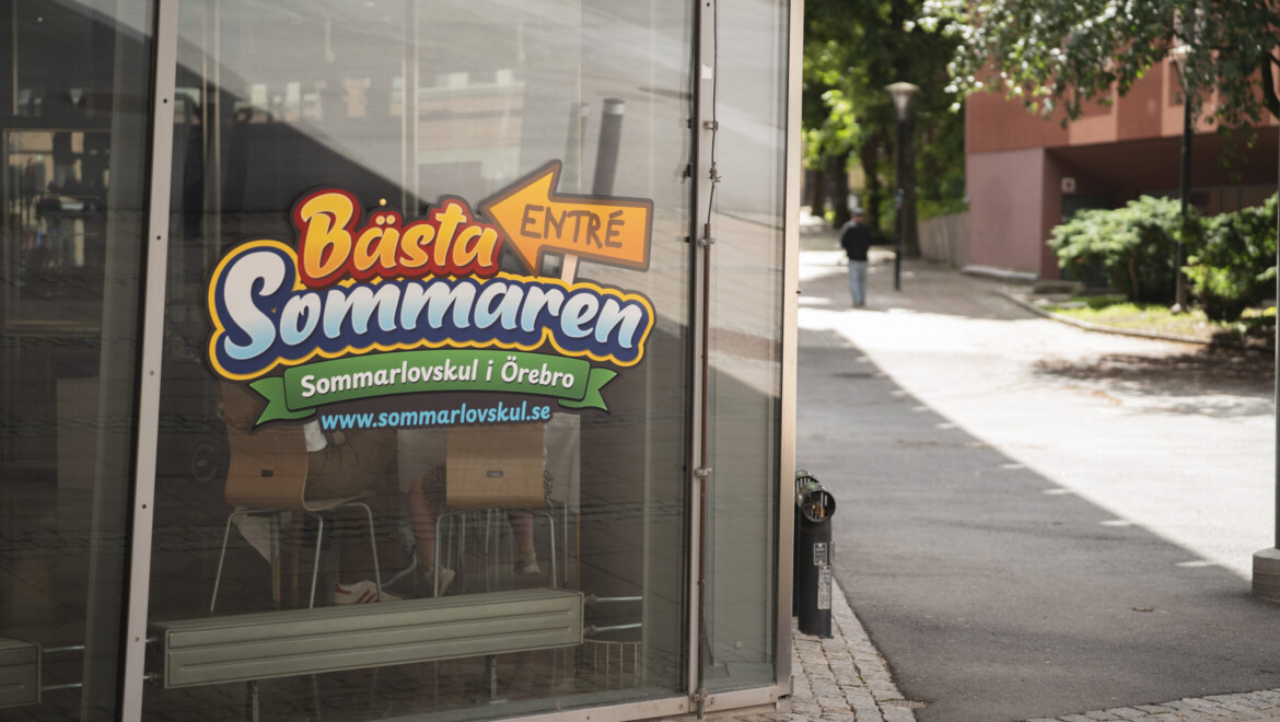 En färgglad logotyp sitter på en glasvägg. Bakom logotypen syns två personer sitta på varsin stol.