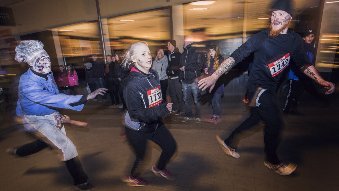 Två löpare blir jagade av en person utklädd till zombie. Personen har kockkläder och är sminkad med sår i ansiktet. Den ena löparen sträcker sig efter den andra. I bakgrunden står flera personer.